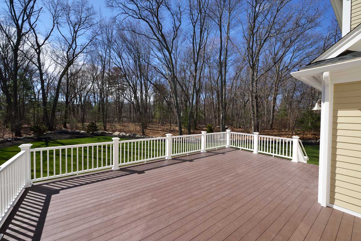 An elevated wooden deck with a white railing looking out to a backyard surrounded by tall bare trees